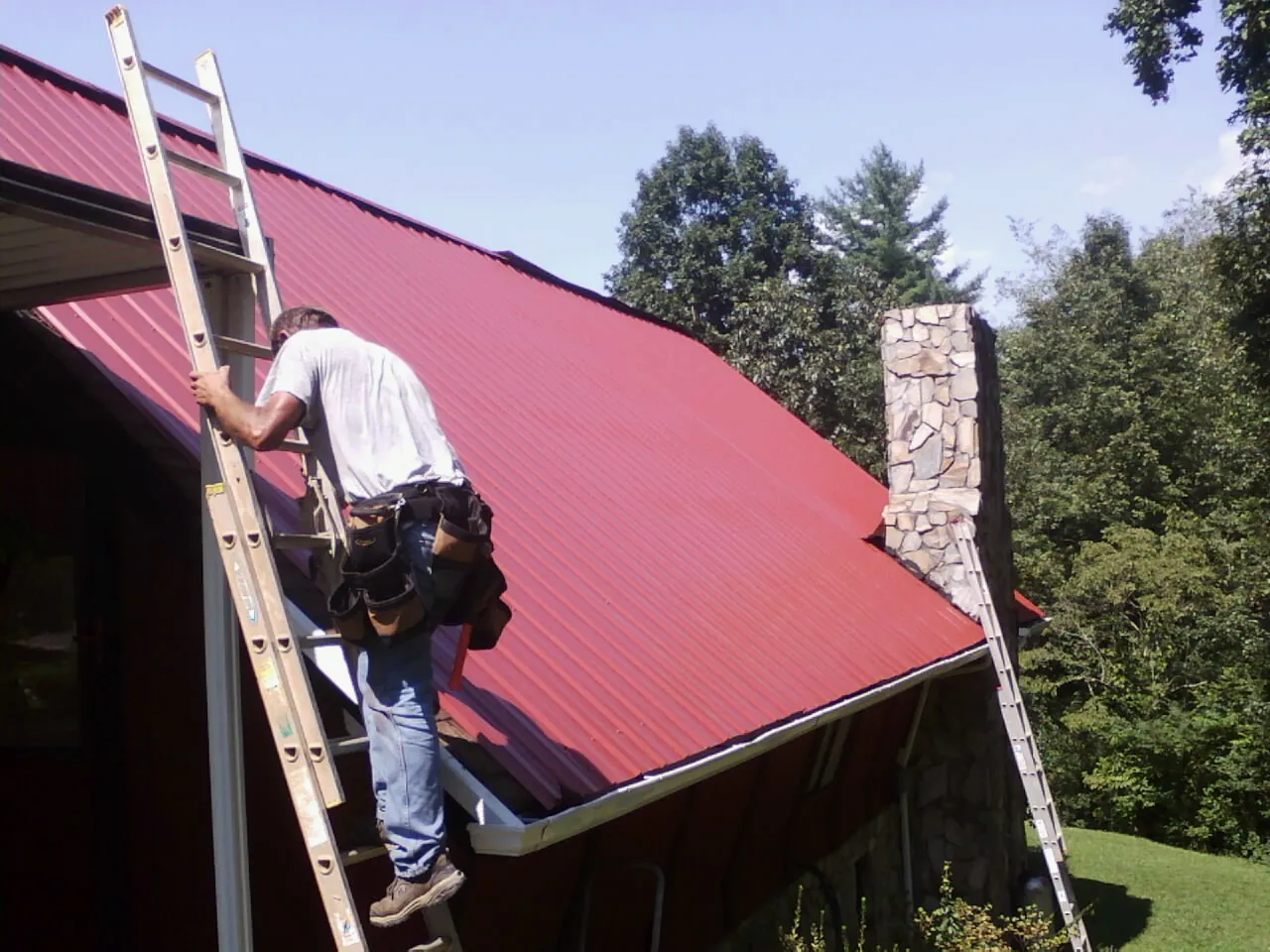 Roofer on a ladder beside a red metal roof and stone chimney — Soffit & Fascia Repair by Roofing Brothers in Buzzards Bay