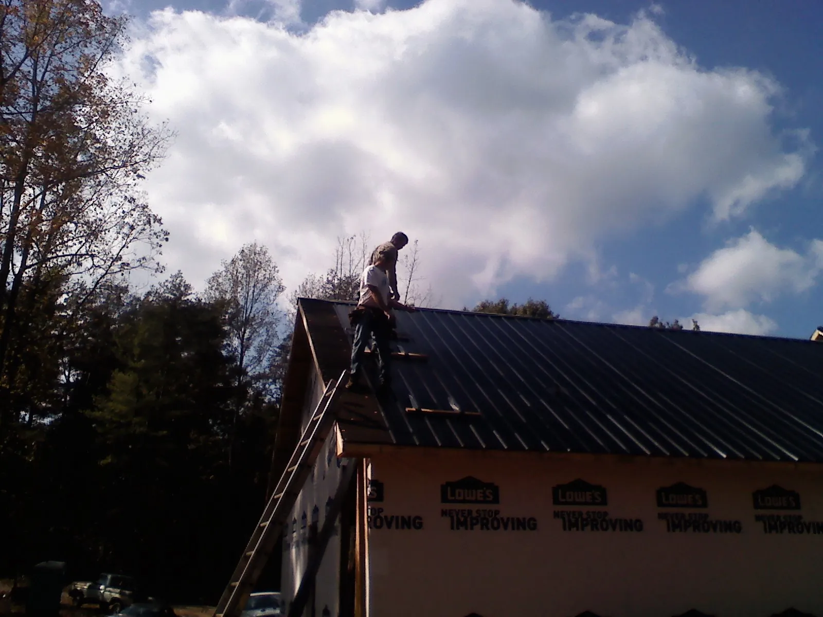 Roofer fastening dark metal panels high against a cloudy sky, from a Roof Inspection job in Buzzards Bay